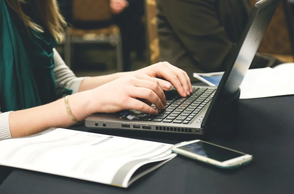 A person typing on a laptop. There is a phone and some paper on the desk.