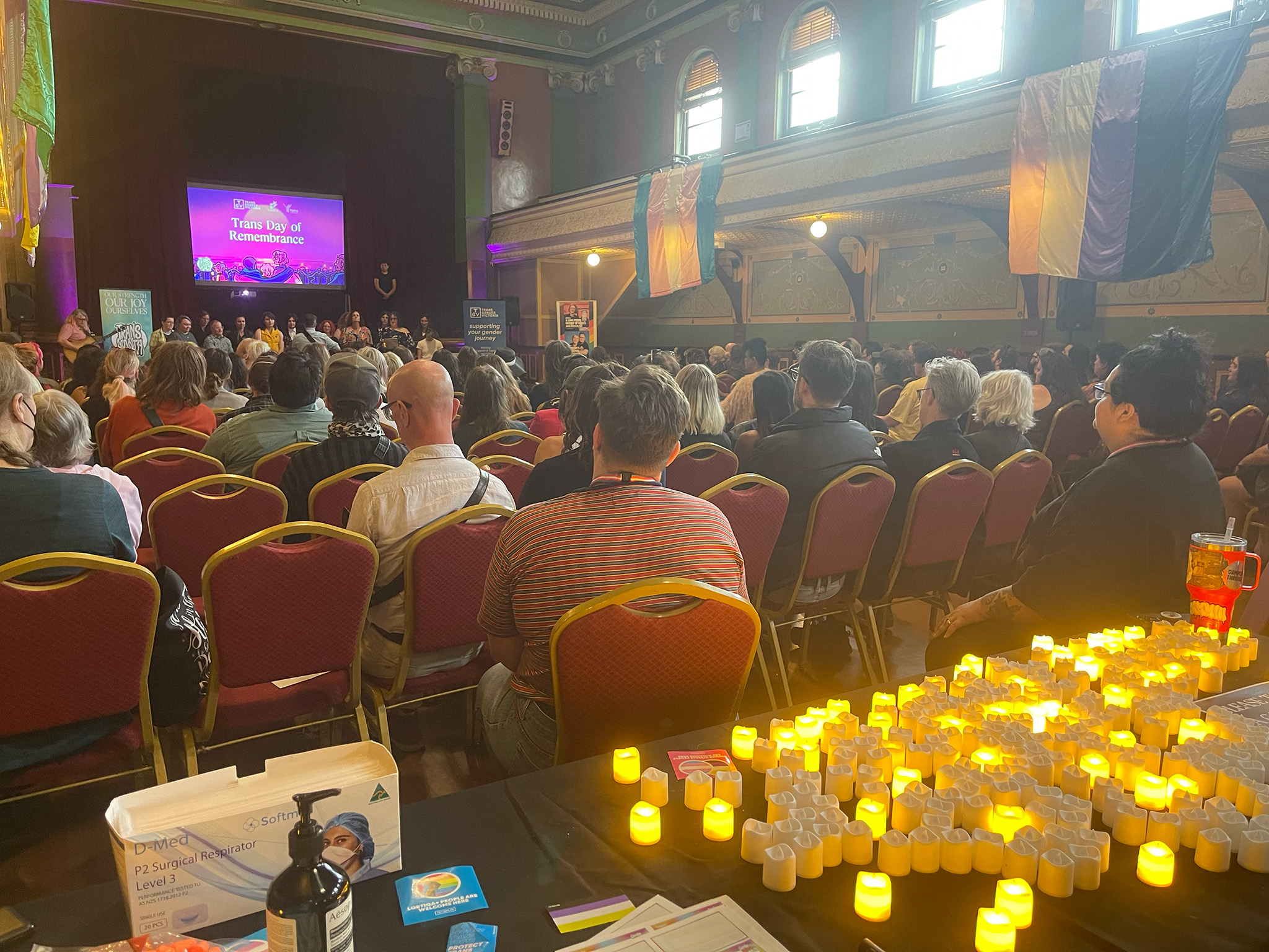 A big hall full of people, facing away from the camera looking towards the stage. In the foreground is a table filled with candles lit up in memory of the loved ones lost to anti-trans violence.