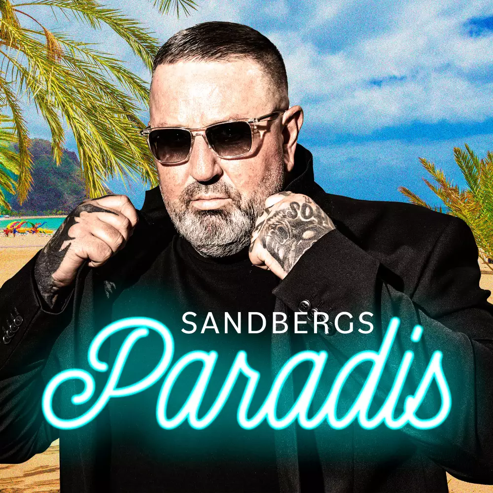 Mand med tatoverede hænder og solbriller justerer sin sorte jakke på en strand med palmer.