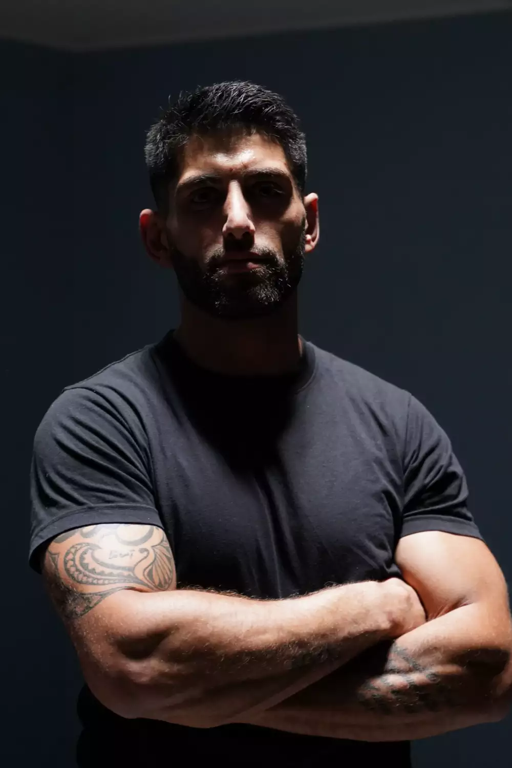 Mand med tatoveringer, korslagte arme og sort t-shirt foran mørk baggrund.