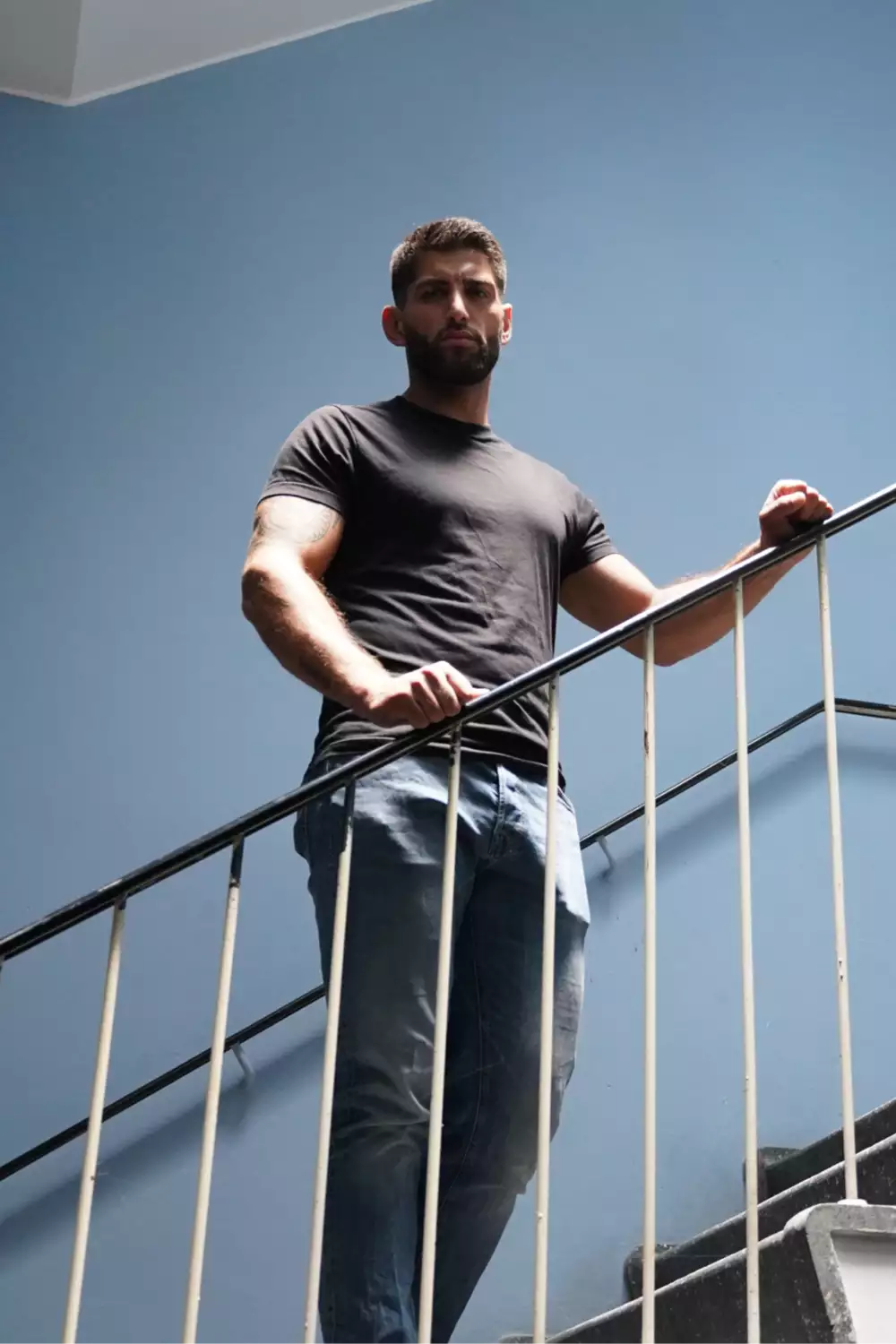 Mand iført sort t-shirt og jeans, står på en trappe med blå væg i baggrunden.