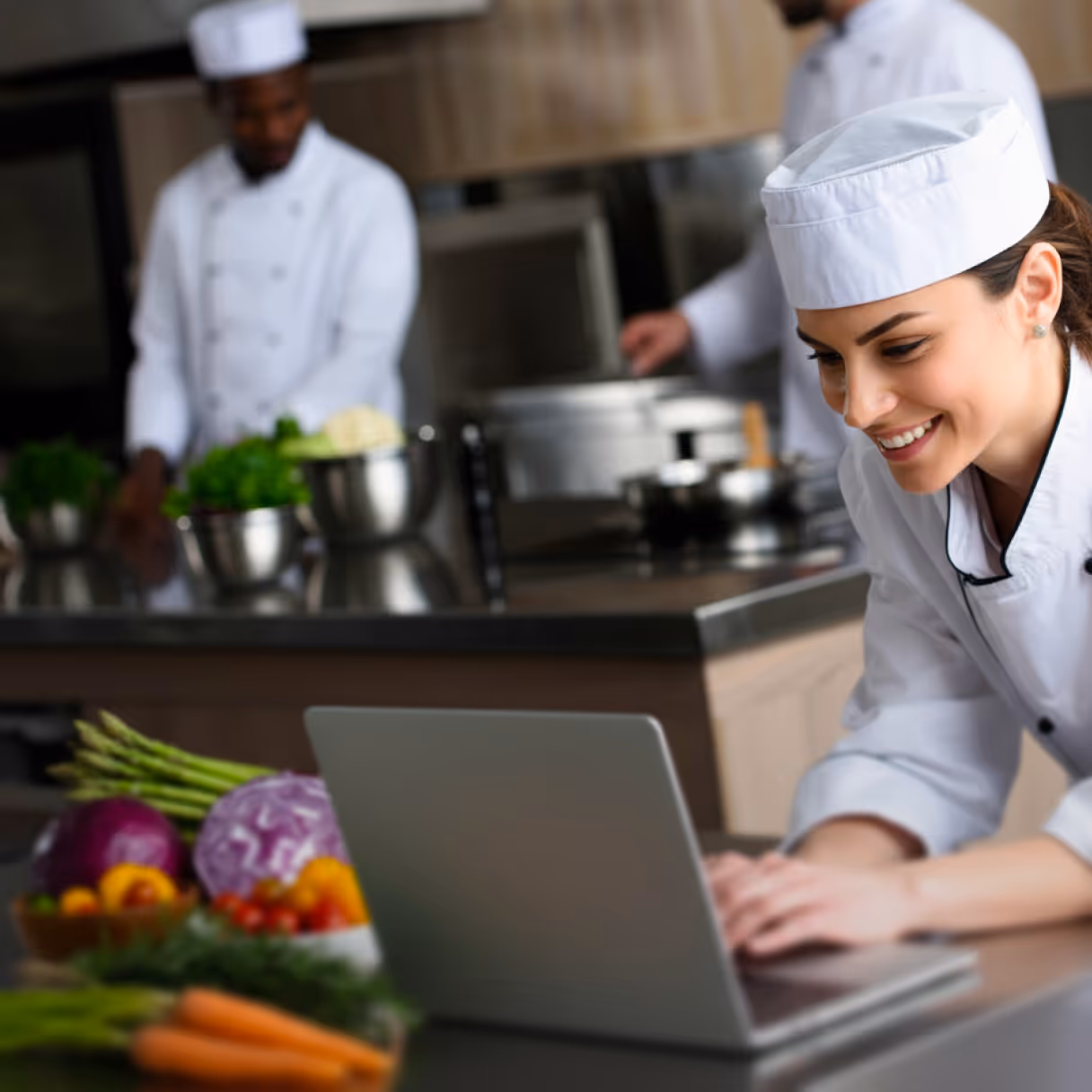 Smiling female chef in white uniform using a laptop in a kitchen with fresh vegetables on the counter.