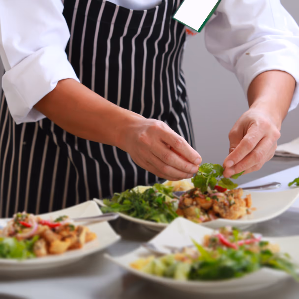 Chef wearing a black and white striped apron garnishing a dish with fresh herbs.