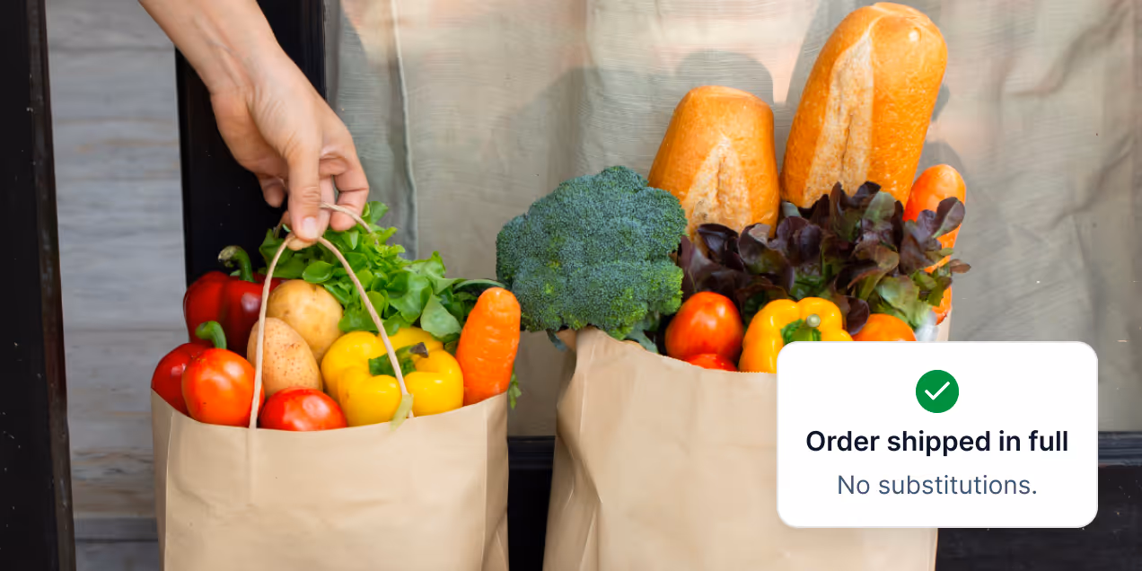 Two paper grocery bags filled with fresh vegetables and bread, with a hand holding one bag and a label stating 'Order shipped in full, No substitutions.'