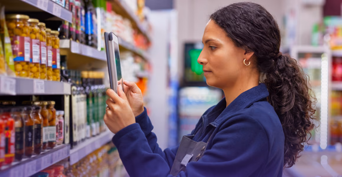 Woman using a tablet to check products on shelves in a grocery store aisle.