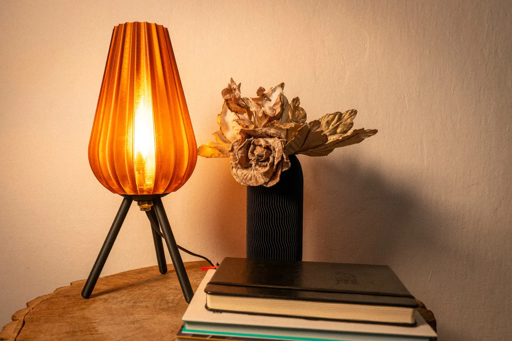 Lampă cu abajur portocaliu și suport negru pe trei picioare, alături de un vas negru cu flori uscate aurii și o grămadă de cărți pe o masă de lemn.