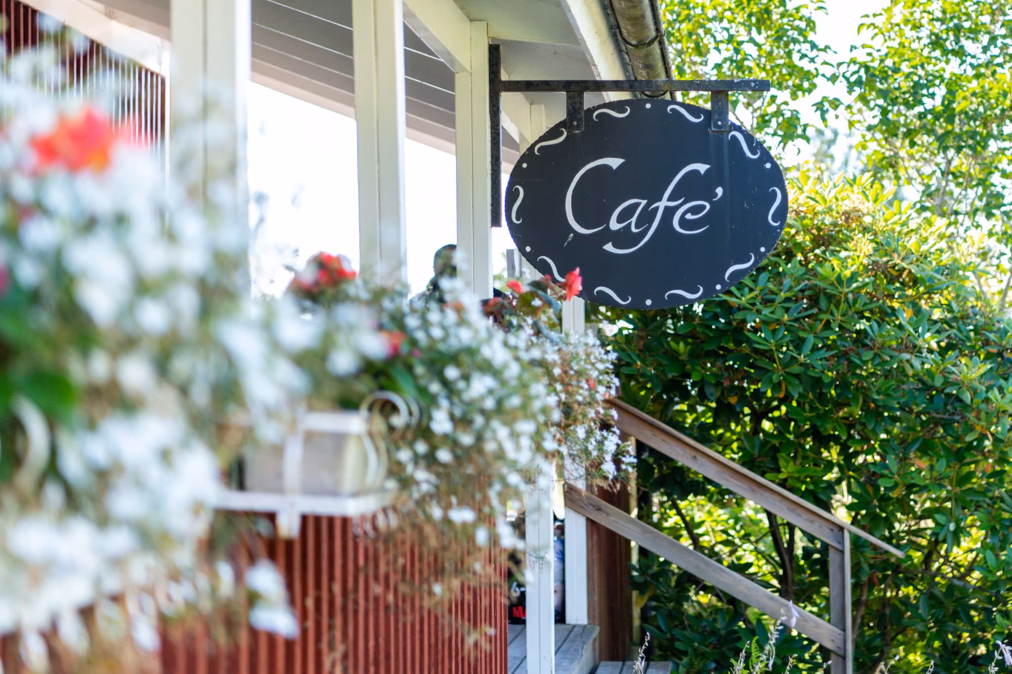 Svart oval skylt med texten ”Café” i vitt som hänger utanför en veranda omgiven av blommor och gröna buskar.