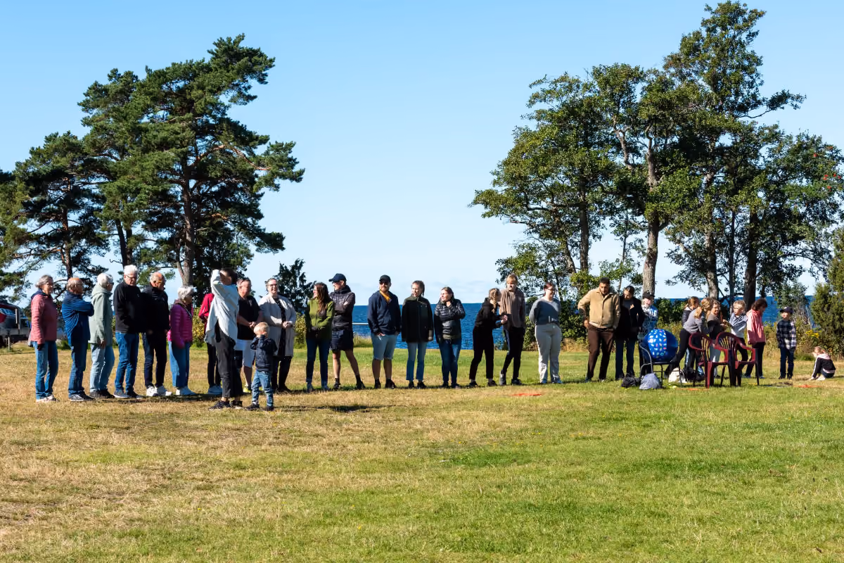Grupp människor som står på en gräsmatta intill träd, med blå himmel och vatten i bakgrunden.