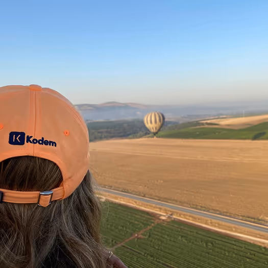 Person wearing an orange Kodem cap observing a striped hot air balloon over fields and hills under a clear blue sky.