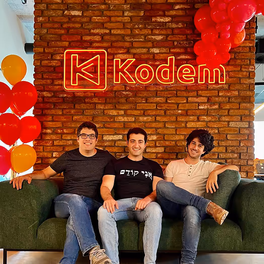 Three smiling men sitting on a green couch under a brick wall with a red neon Kodem sign and red and orange balloons on the left.