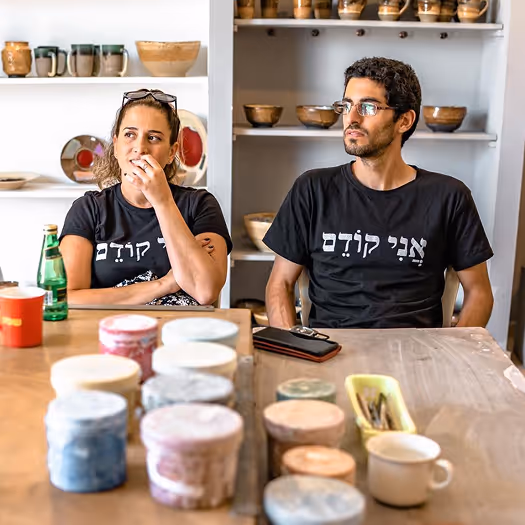 Two people wearing black shirts with Hebrew text sitting at a wooden table with pottery tools and ceramic containers, with shelves of pottery in the background.