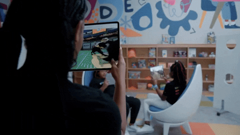 Person holding a tablet showing a virtual tennis game while two children sit in a classroom with colorful wall art and bookshelves.