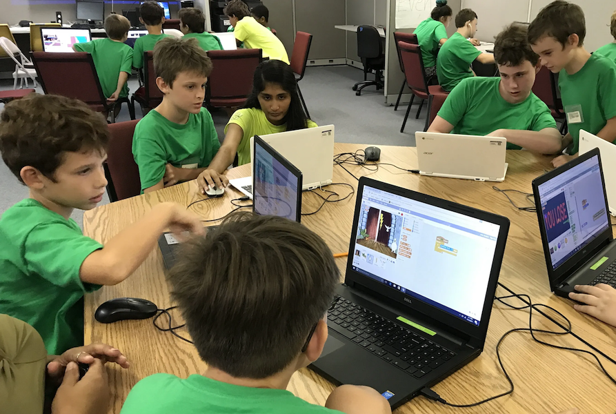 A group of children wearing green and yellow shirts working on laptops around a large table in a classroom setting.