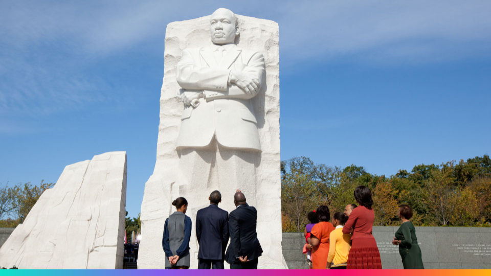 Visitors standing and looking up at the large white marble statue of Dr. Martin Luther King Jr. with arms crossed under a blue sky.
