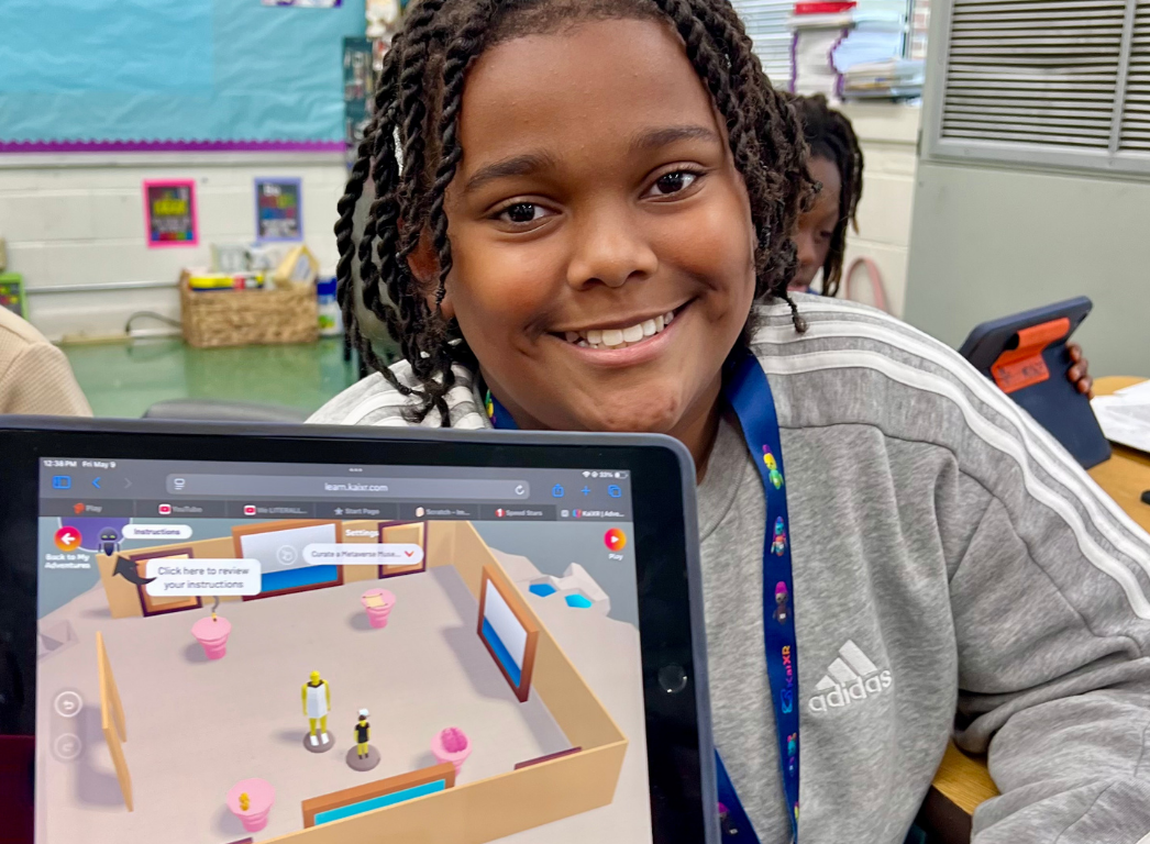 Five smiling students sitting at desks in a classroom, each with a tablet showing a virtual environment on screen.