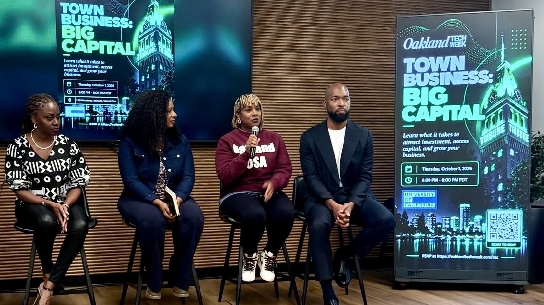 Four panelists seated on stools in front of a wood-paneled wall during Oakland Tech Week's Town Business: Big Capital event, with a large digital sign displaying event details to the right.