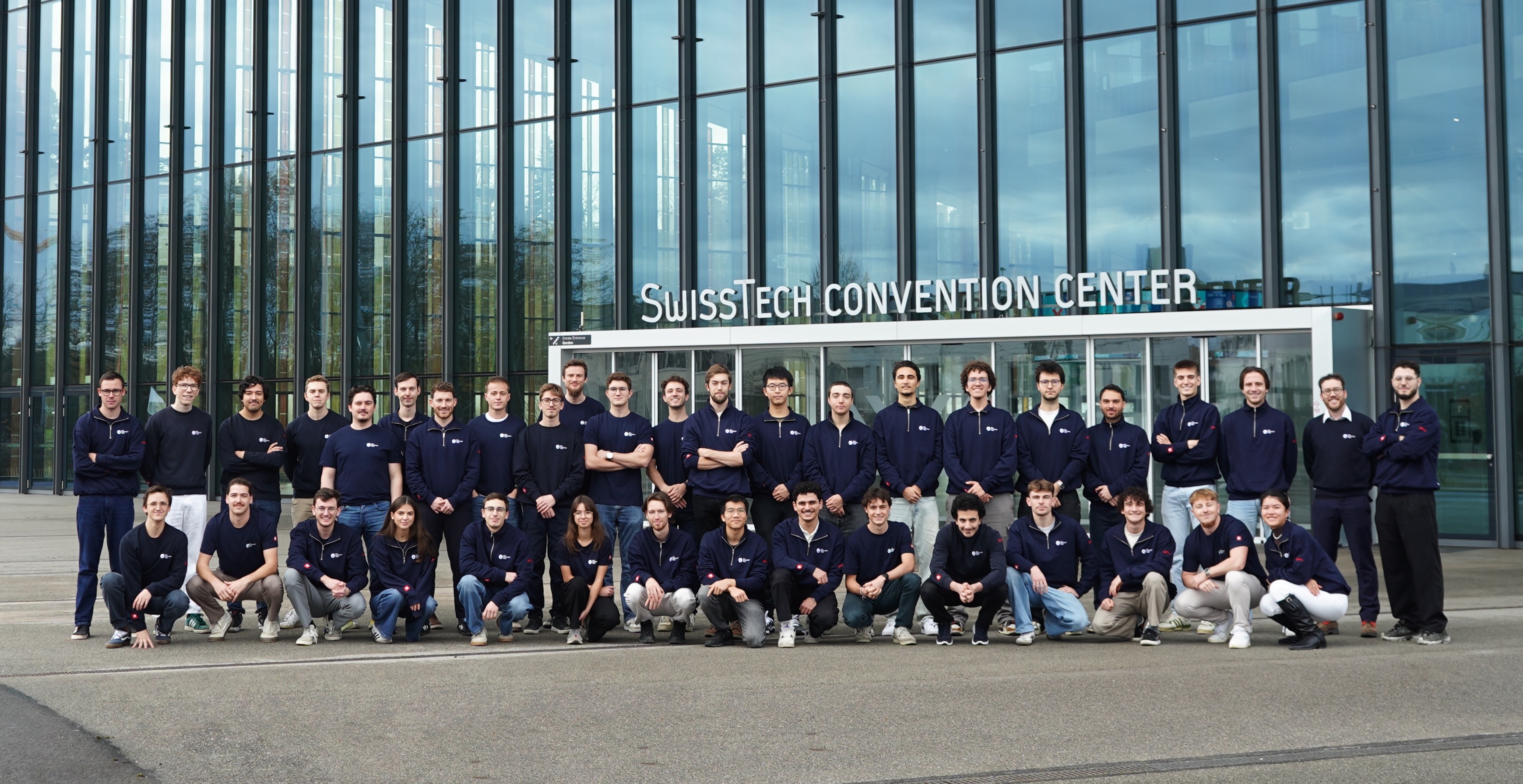 Group of young adults in black t-shirts posing outside the Swiss Tech Convention Center with glass facade.