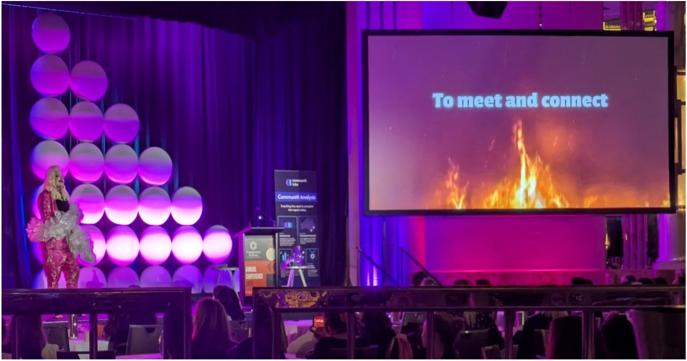 A speaker wearing a pink and white costume stands on stage at a conference with a purple-lit backdrop of circular panels. A large screen beside them displays the words “To meet and…” with flames in the background, while an audience watches from their tables.