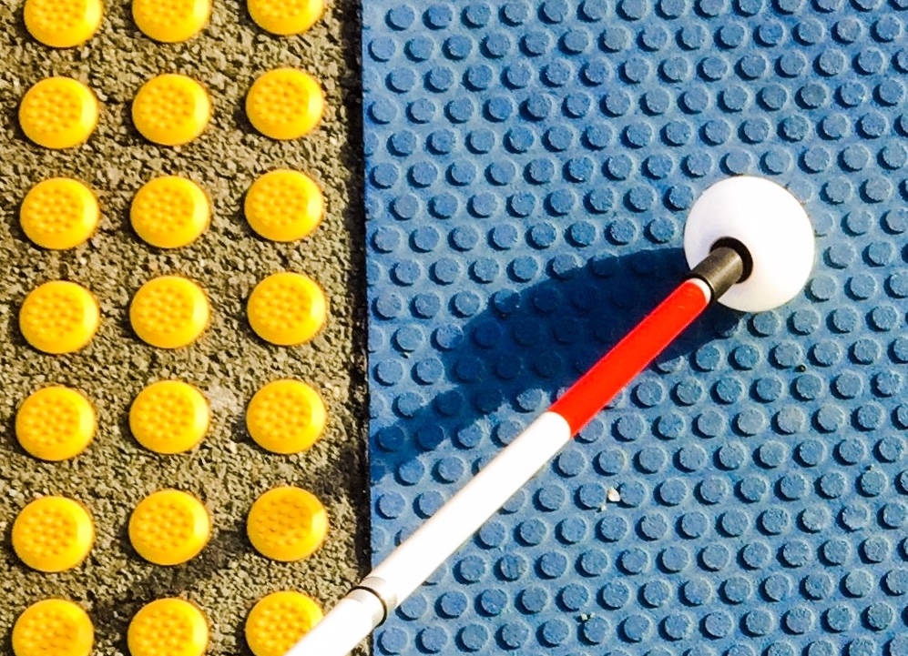 Photo of a white cane touching tactile ground surface indicators on a paved footpath — yellow blister warning tiles at a crossing.
