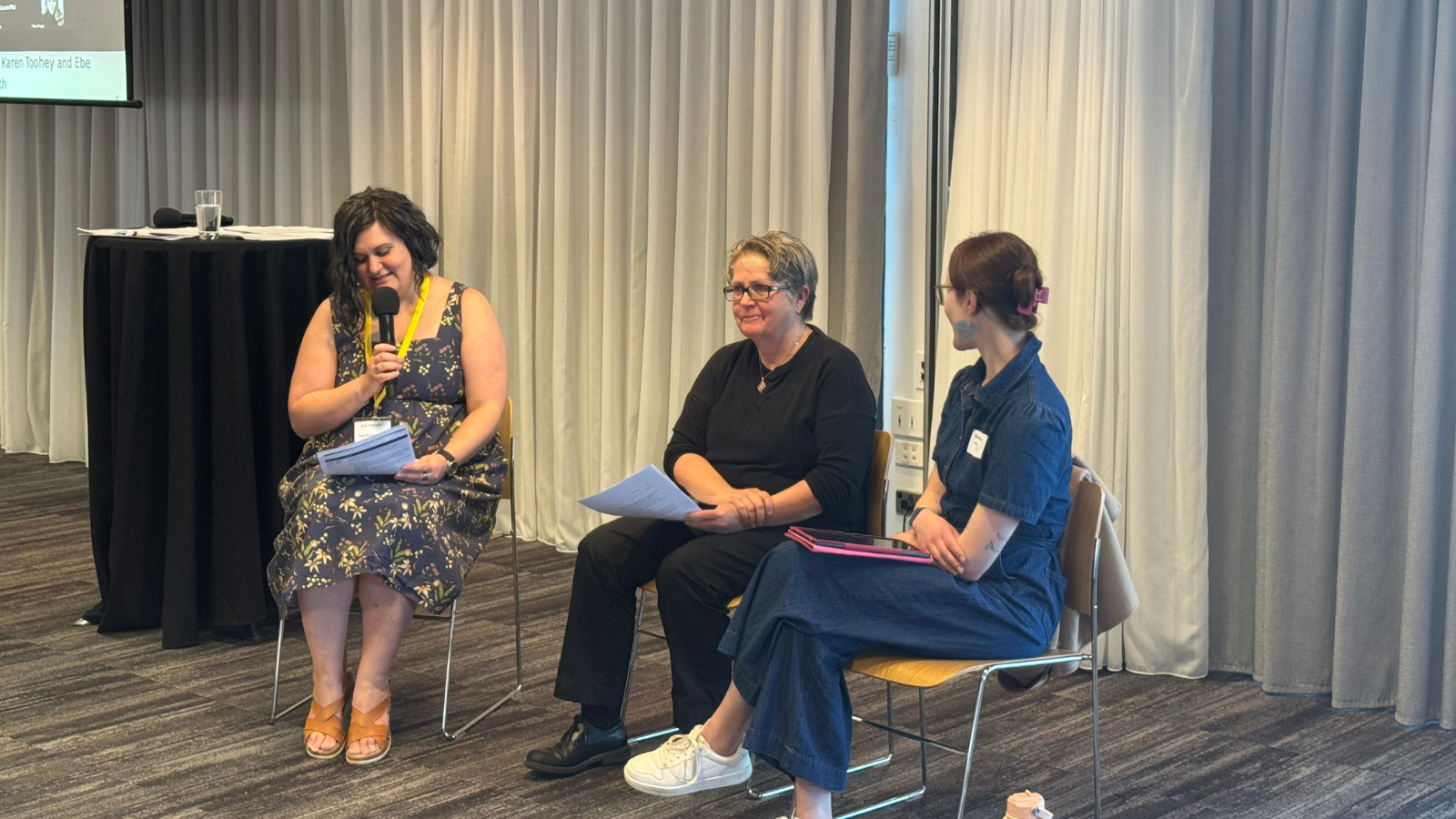 Three people seated in chairs at a panel discussion event, with one holding a microphone and notes.