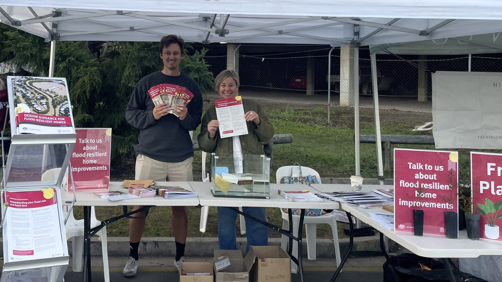 Two people smile at a community stall under a marquee, holding flood resilience brochures and fact sheets. A sign reads "Talk to us about flood resilient home improvements.