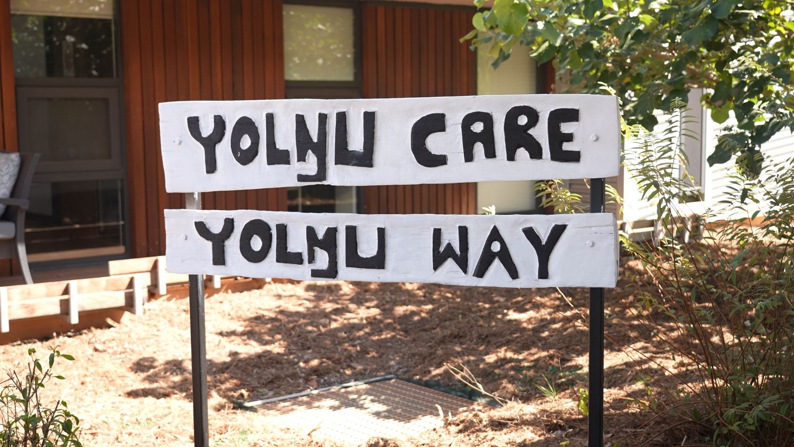 A hand-painted wooden sign reading "Yolŋu Care Yolŋu Way" in front of a timber building surrounded by native garden plantings.