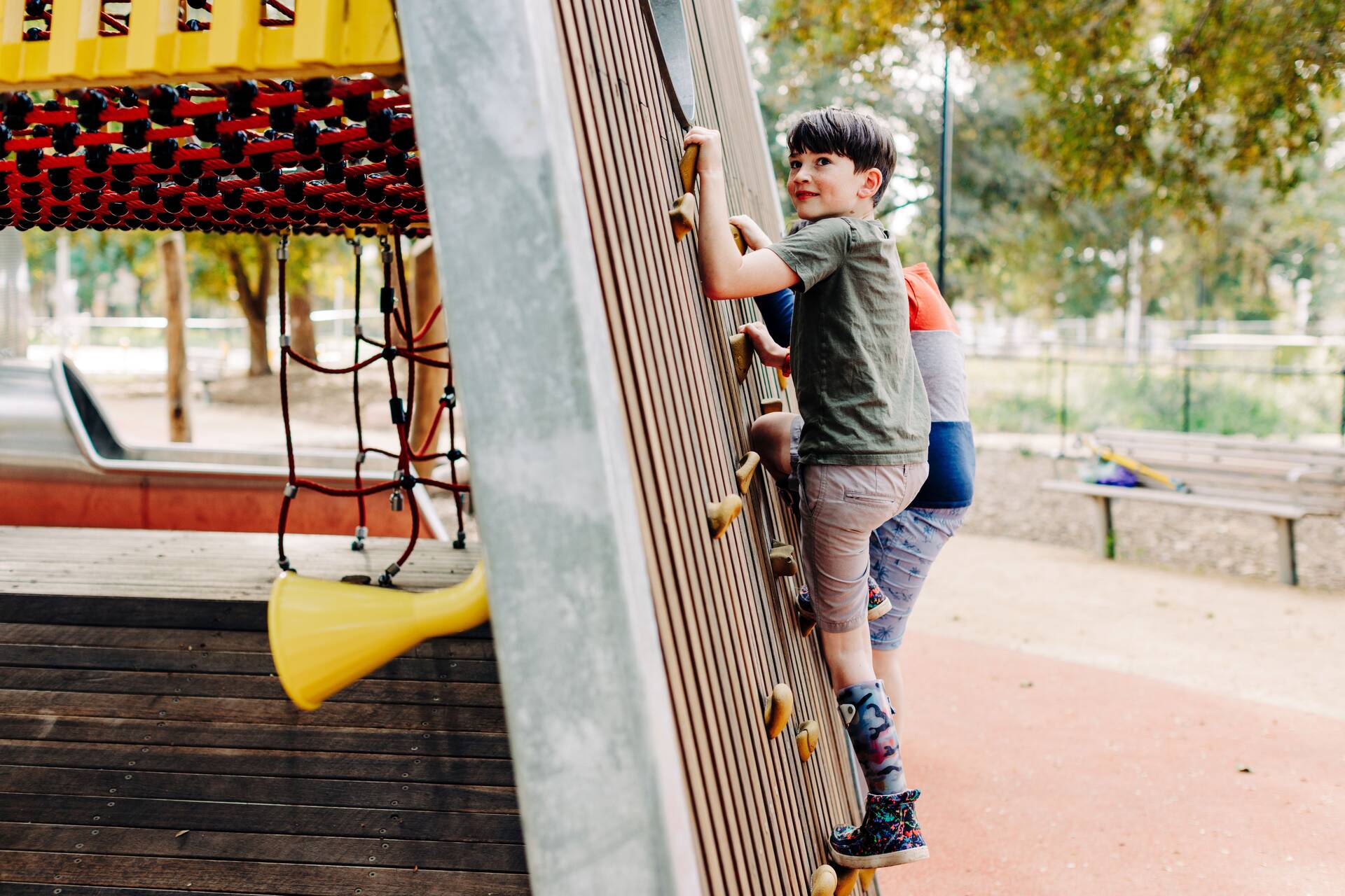 A child climbs a wooden wall on a playground structure, wearing a green t-shirt and a leg brace. Another child is visible behind them. Play equipment and trees are in the background.