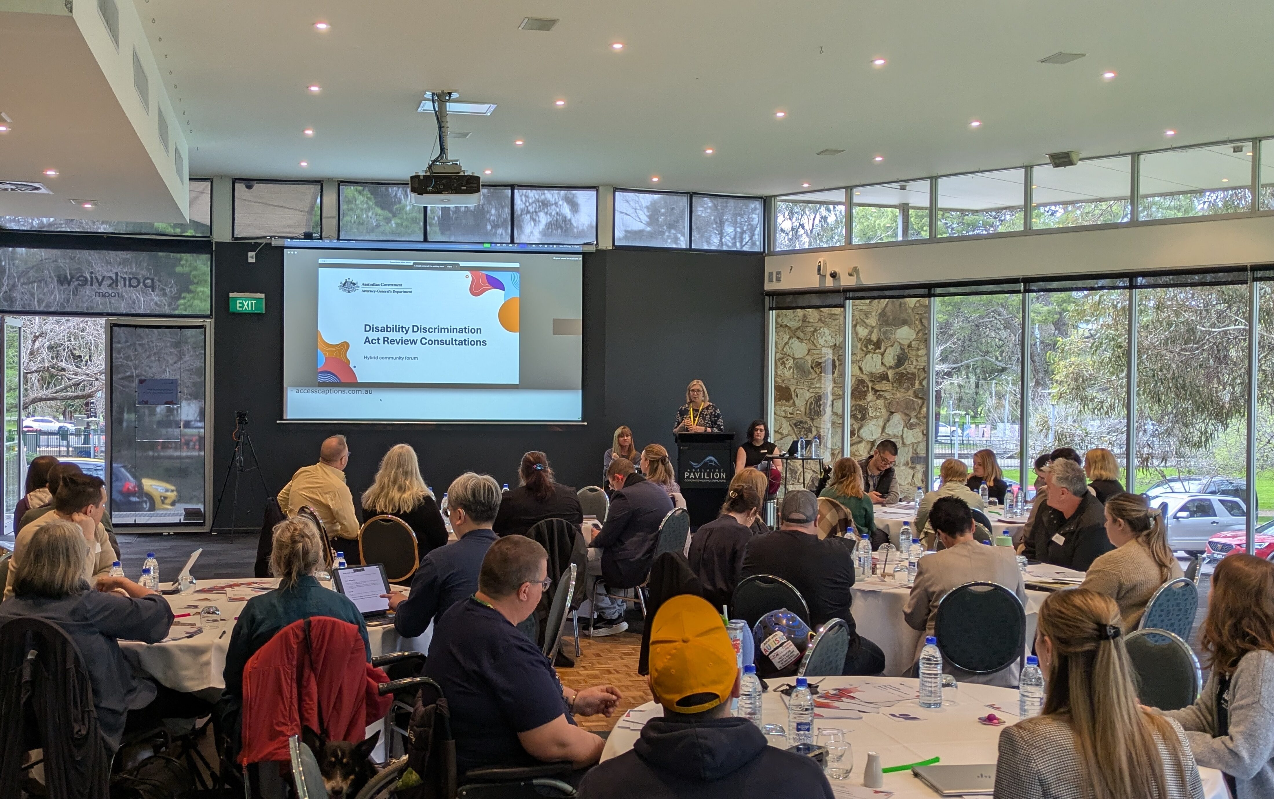 Participants seated at round tables during the Disability Discrimination Act Review Consultations hybrid community forum, with a presenter at the podium and the session title displayed on a projector screen.