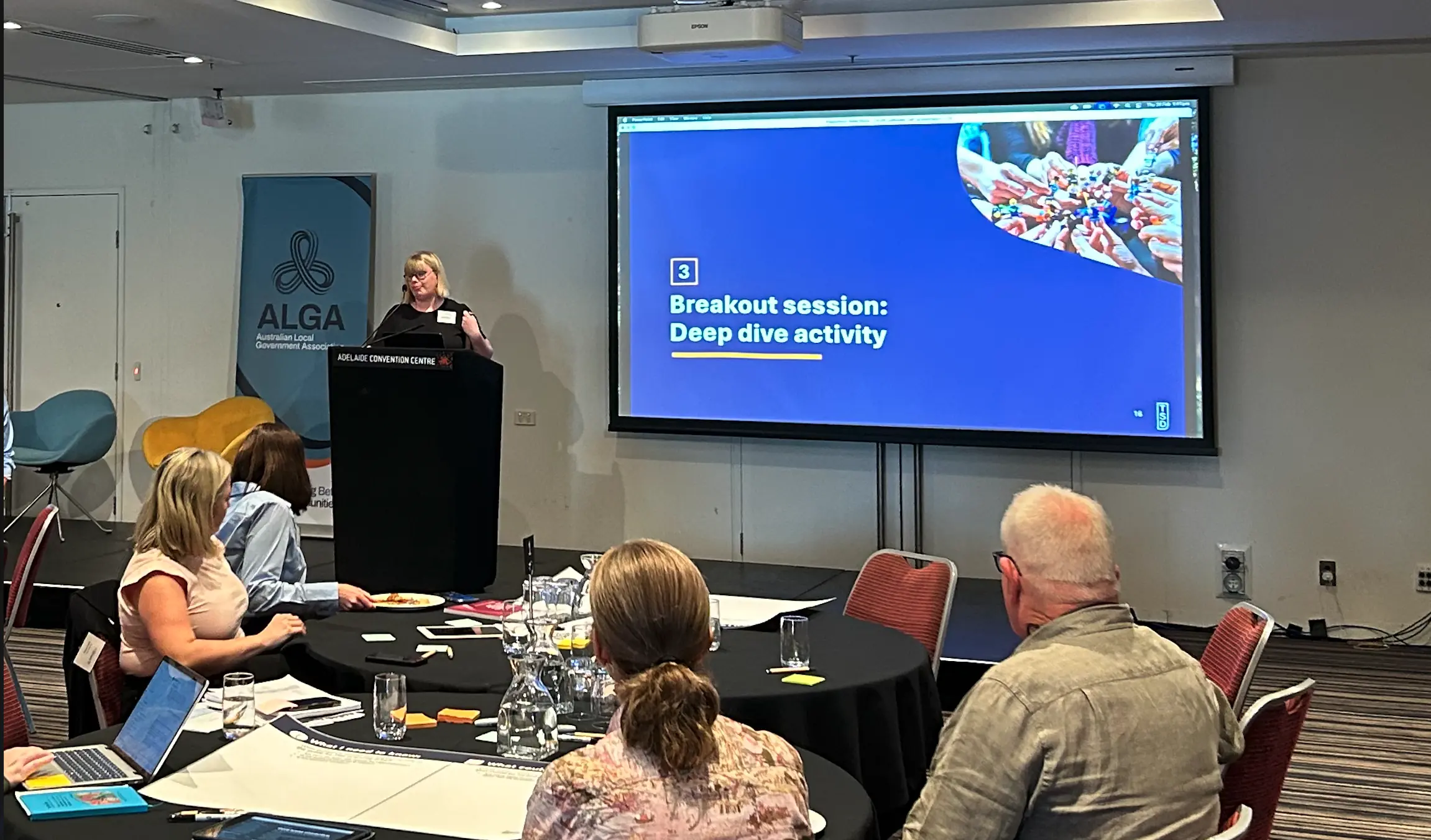 A facilitator stands at a podium in front of a large screen reading "Breakout session: Deep dive activity" with an ALGA banner beside her, while attendees seated at round tables look on.