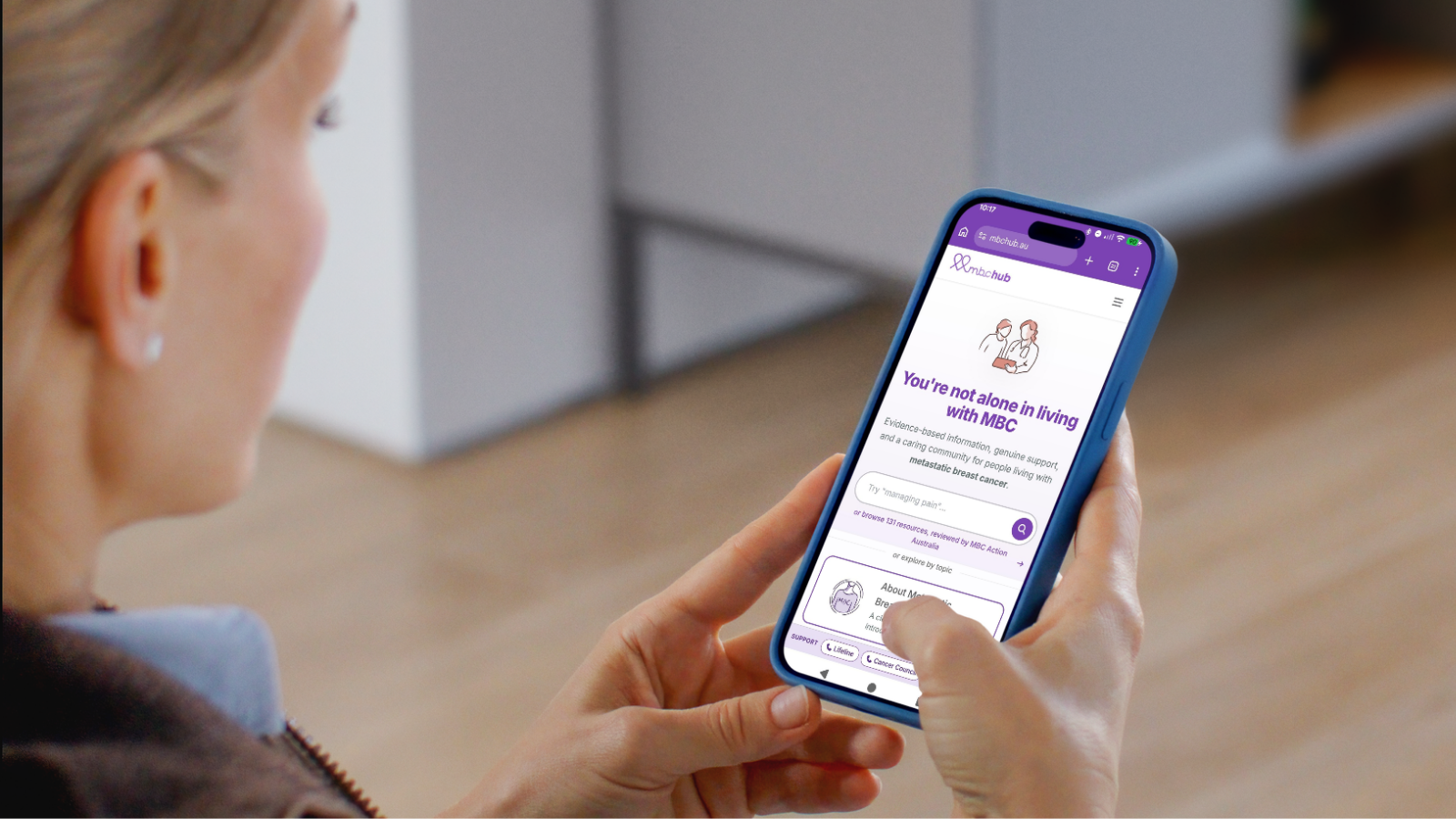 A woman looks at her phone displaying the MBC Hub website homepage, with the heading "You're not alone in living with MBC" and a search bar to browse resources for people living with metastatic breast cancer.