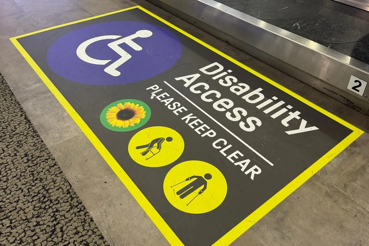 Photo of a sign on the ground in front of a baggage carousel. The sign has a large disability icon and smaller icons of a sunflower, a figure with a walking frame and a figure with crutches. Text says, “Disability Access. Please keep clear”.