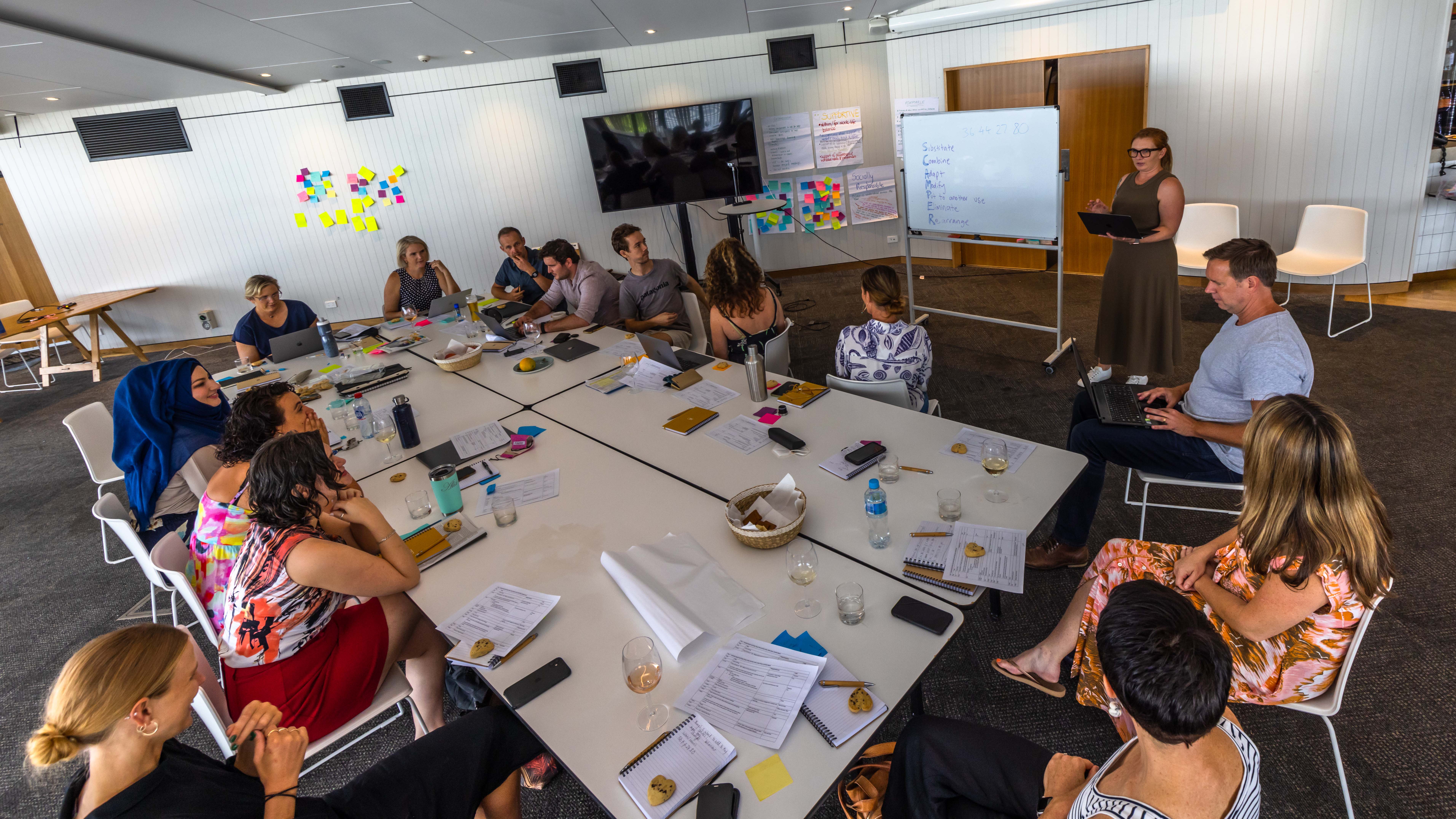 A presenter standing in front of a whiteboard, in front of a group of people sitting around a table. 