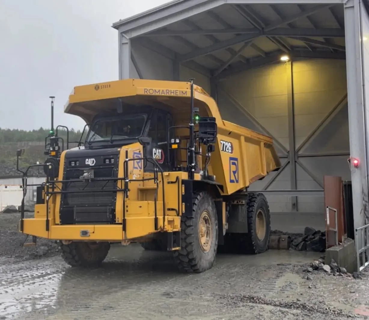 Large yellow and black Caterpillar 772G heavy dump truck parked inside an industrial shelter on a muddy surface.