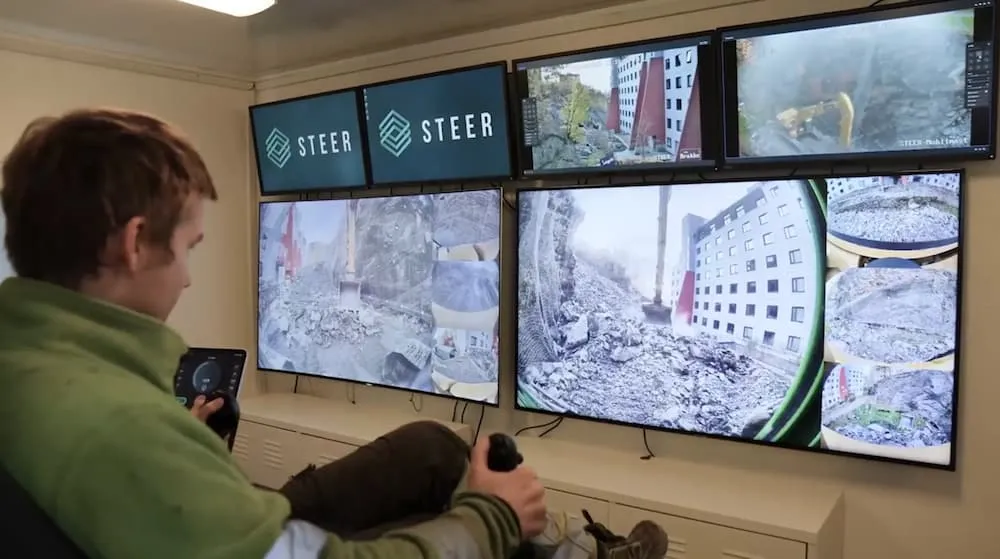Operator using a remote control station with multiple monitors to safely maneuver heavy equipment at a hazardous rockslide site.