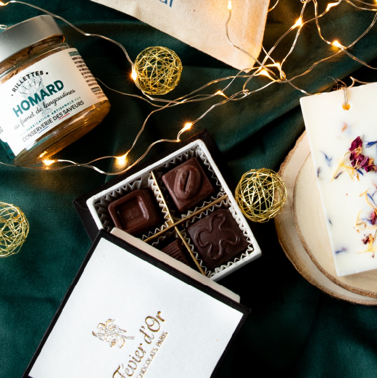 Box cadeau d'entreprise Einaï : chocolats Févier d'Or, rillettes de homard artisanales et tablette parfumée aux fleurs séchées sur un velours vert, entre des guirlandes lumineuses