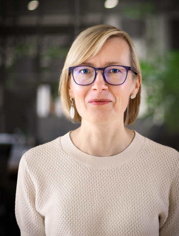 Irina, with shoulder-length blonde hair and wearing a green blouse, poses with a gentle smile. An office is visible in the background