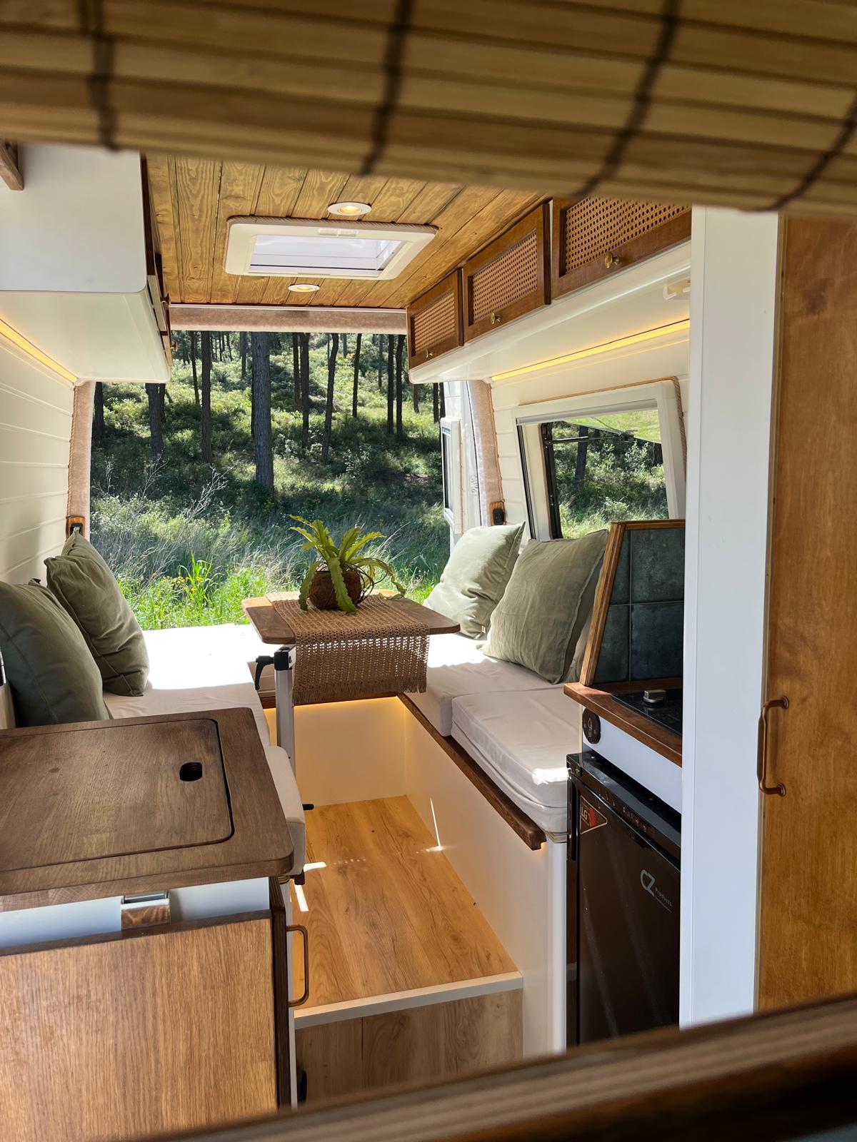 Cozy camper van interior with wooden ceiling, cushioned seating, a small table with a plant, and large windows showing forest greenery outside.