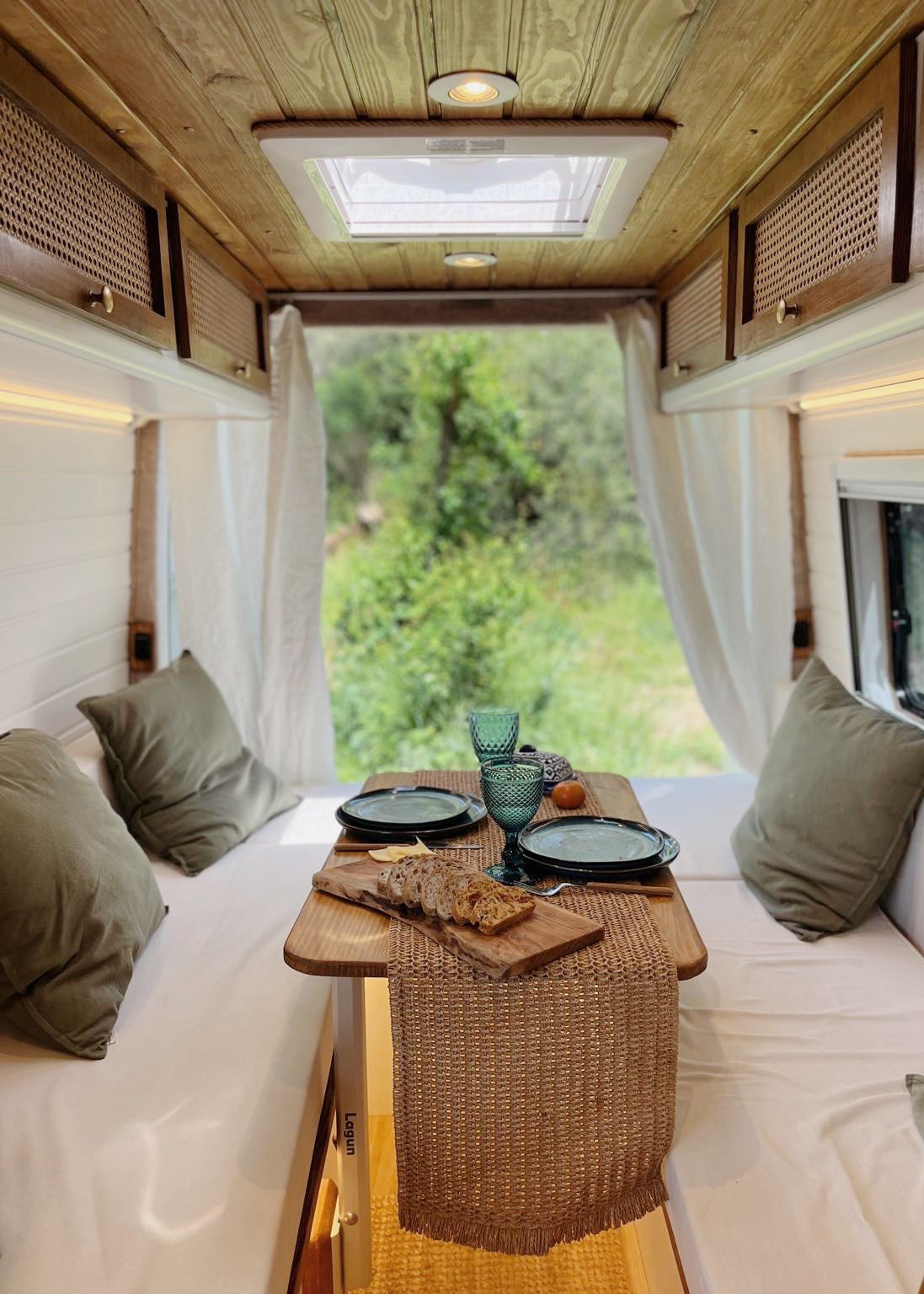 Cozy camper van dining area with wooden table, green cushions, sliced bread on a cutting board, two plates, and green glasses, with greenery visible outside.
