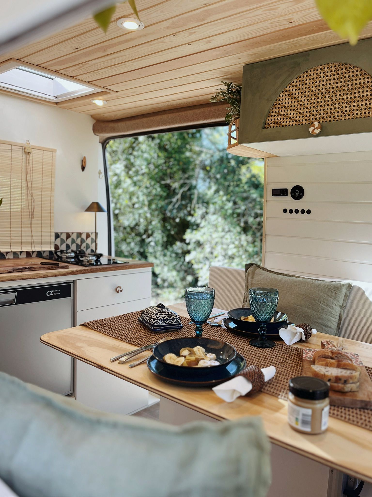 Interior of a wooden-themed camper van with a skylight, a green potted plant on a wooden table, cushioned benches, and a kitchen sink next to an open door showing grass outside.