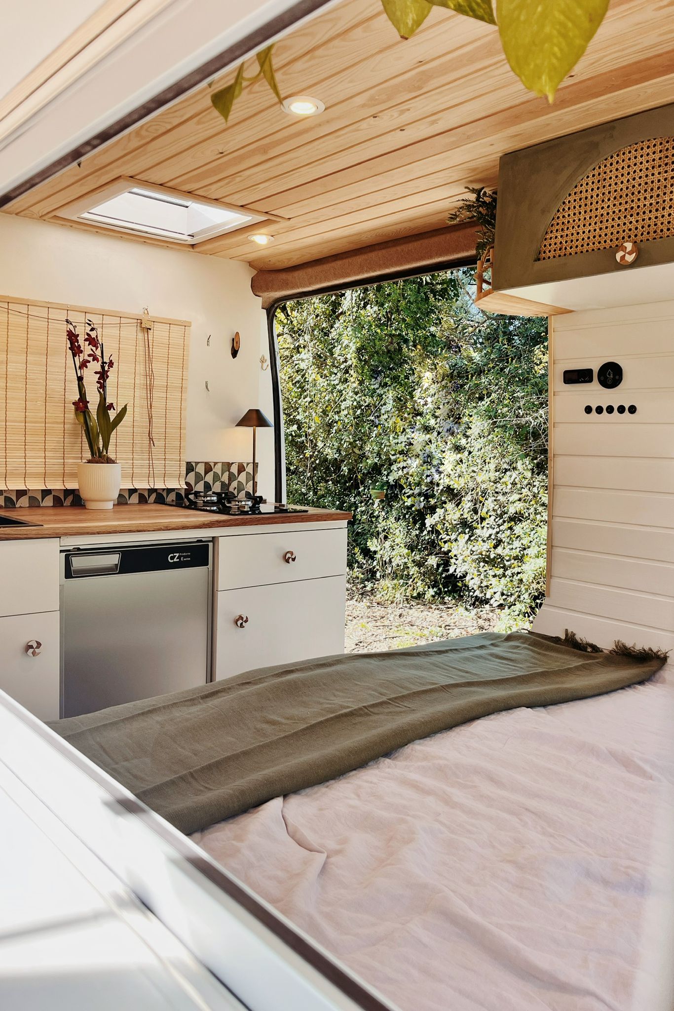 Cozy camper van interior with wooden ceiling, cushioned seating, a small table with a plant, and large windows showing forest greenery outside.