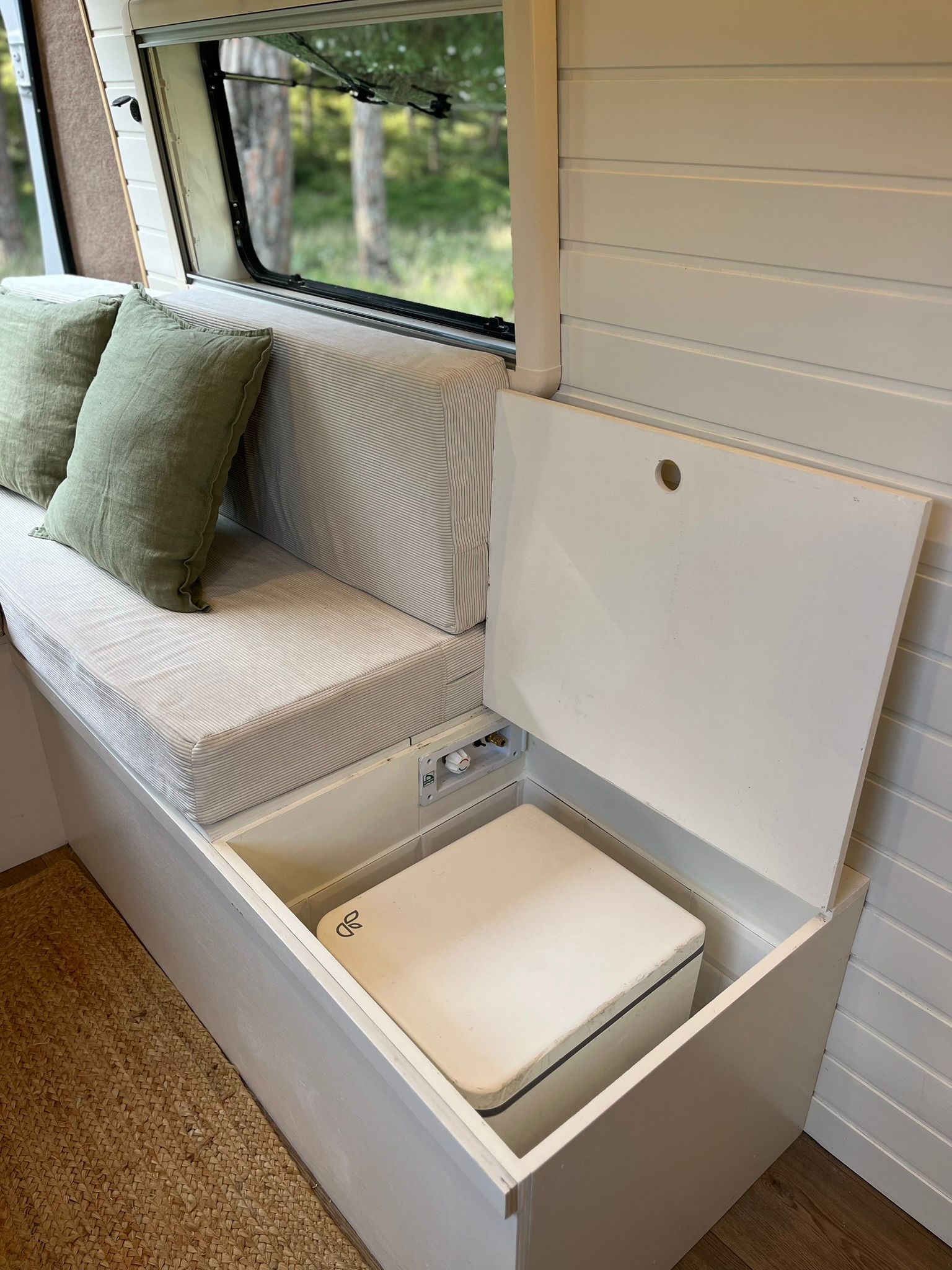 Cozy camper van interior with wooden ceiling, cushioned seating, a small table with a plant, and large windows showing forest greenery outside.