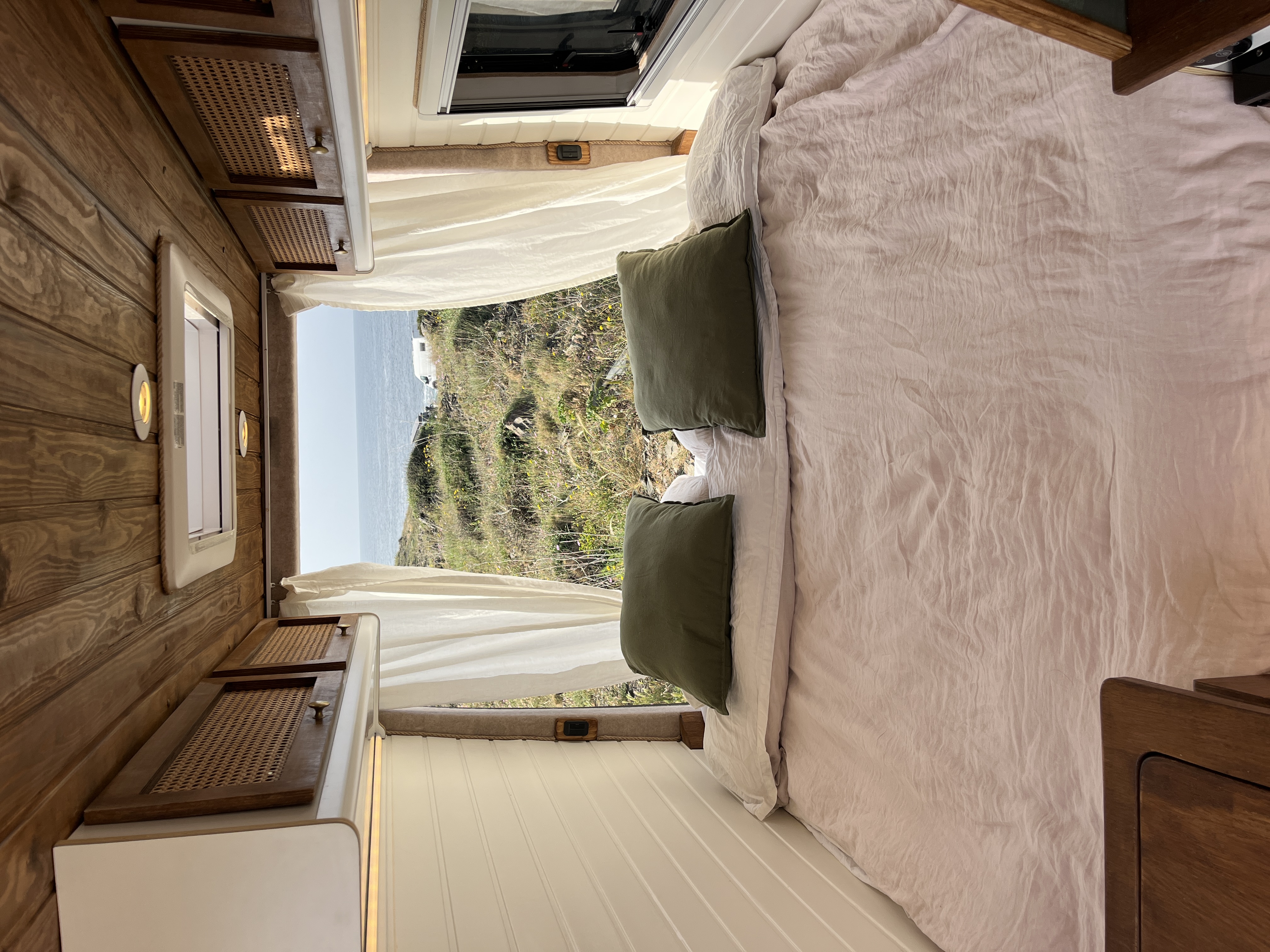 Cozy camper van interior with wooden ceiling, cushioned seating, a small table with a plant, and large windows showing forest greenery outside.