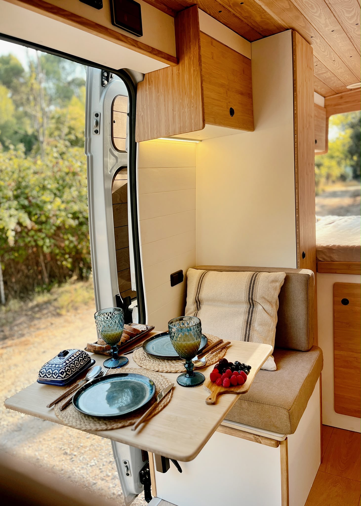 Cozy camper van interior with wooden ceiling, cushioned seating, a small table with a plant, and large windows showing forest greenery outside.