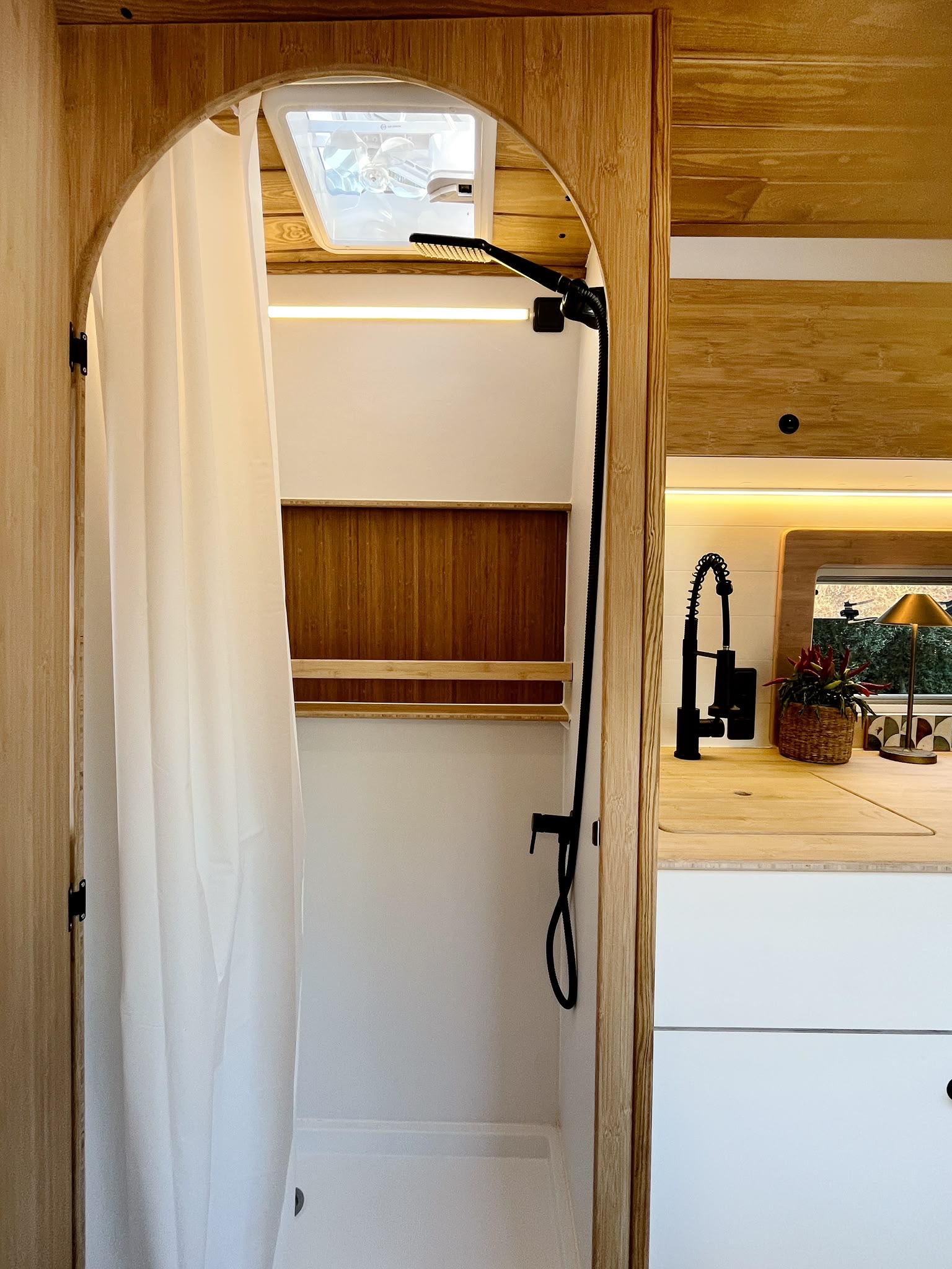 Interior of a compact camper kitchen with wooden finishes, a stove, and a tall wooden door revealing a small storage area inside.