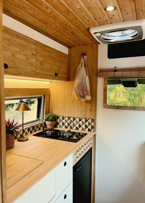Cozy camper van dining area with wooden table, green cushions, sliced bread on a cutting board, two plates, and green glasses, with greenery visible outside.