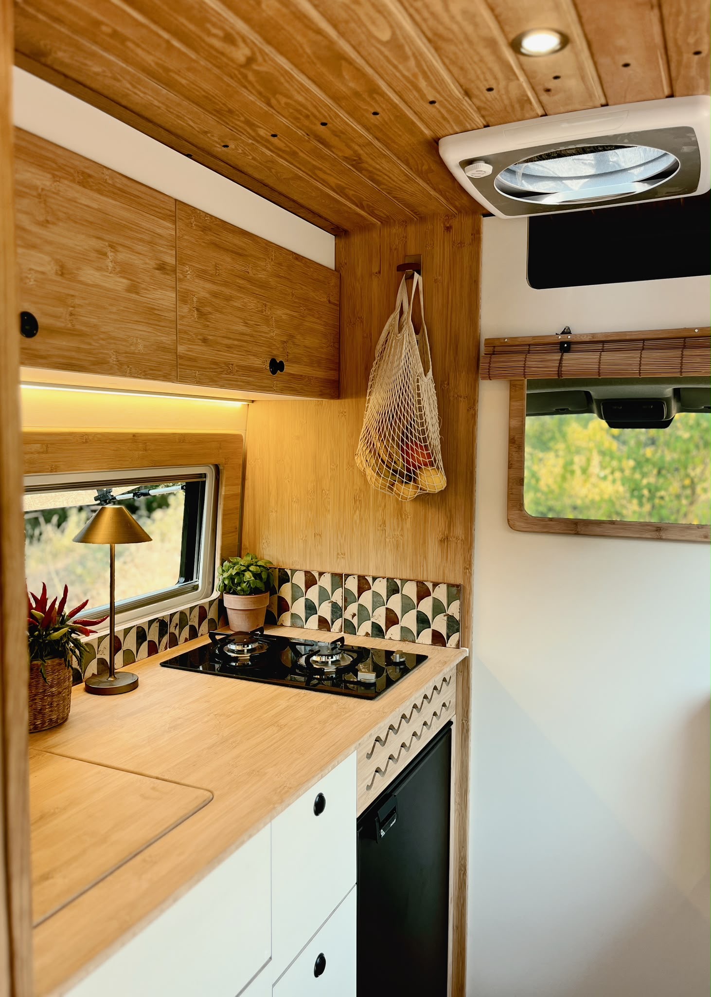 Cozy camper van dining area with wooden table, green cushions, sliced bread on a cutting board, two plates, and green glasses, with greenery visible outside.