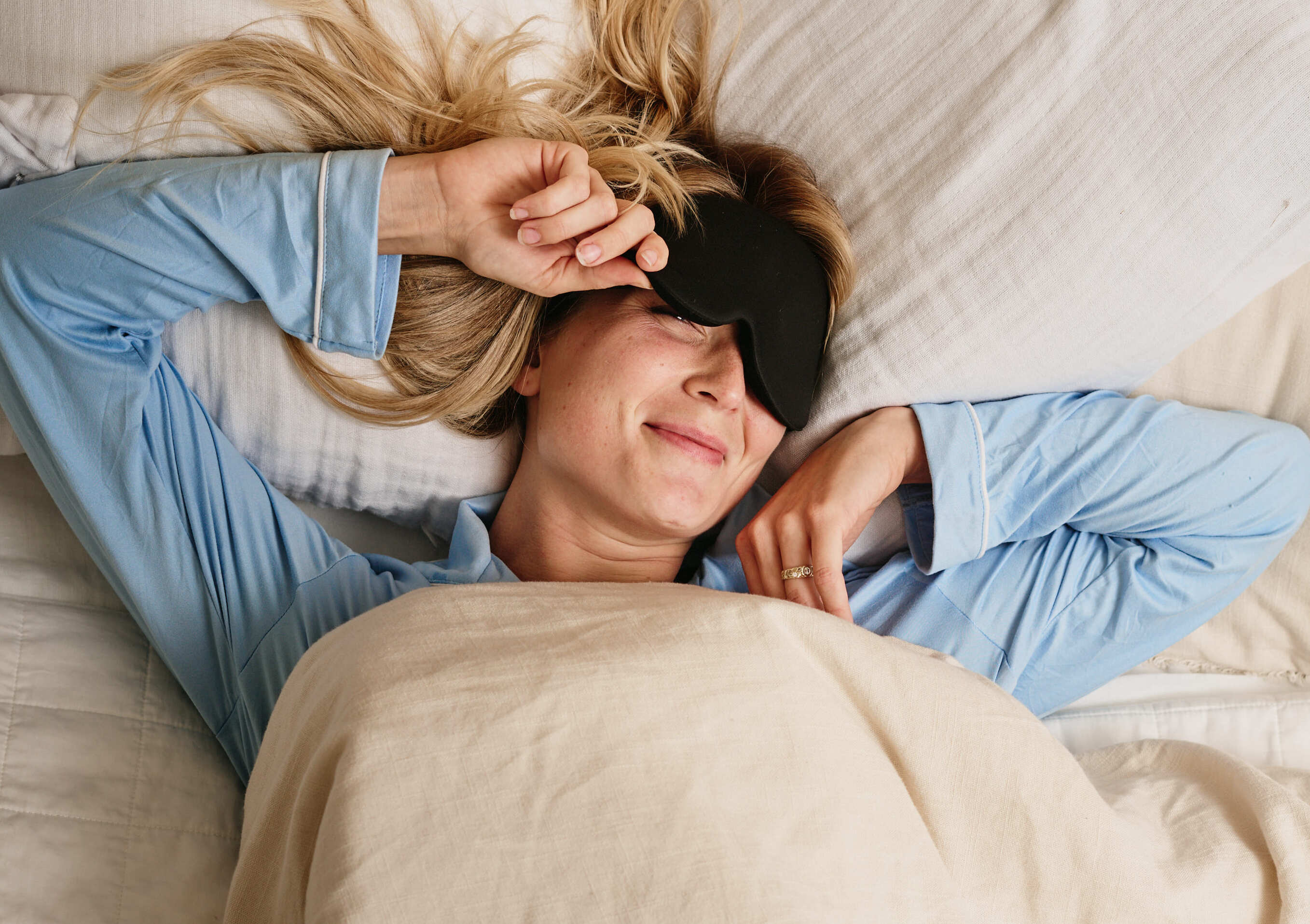 A person sits up in bed, removes a sleep mask from their eyes, and smiles as morning light fills the room.