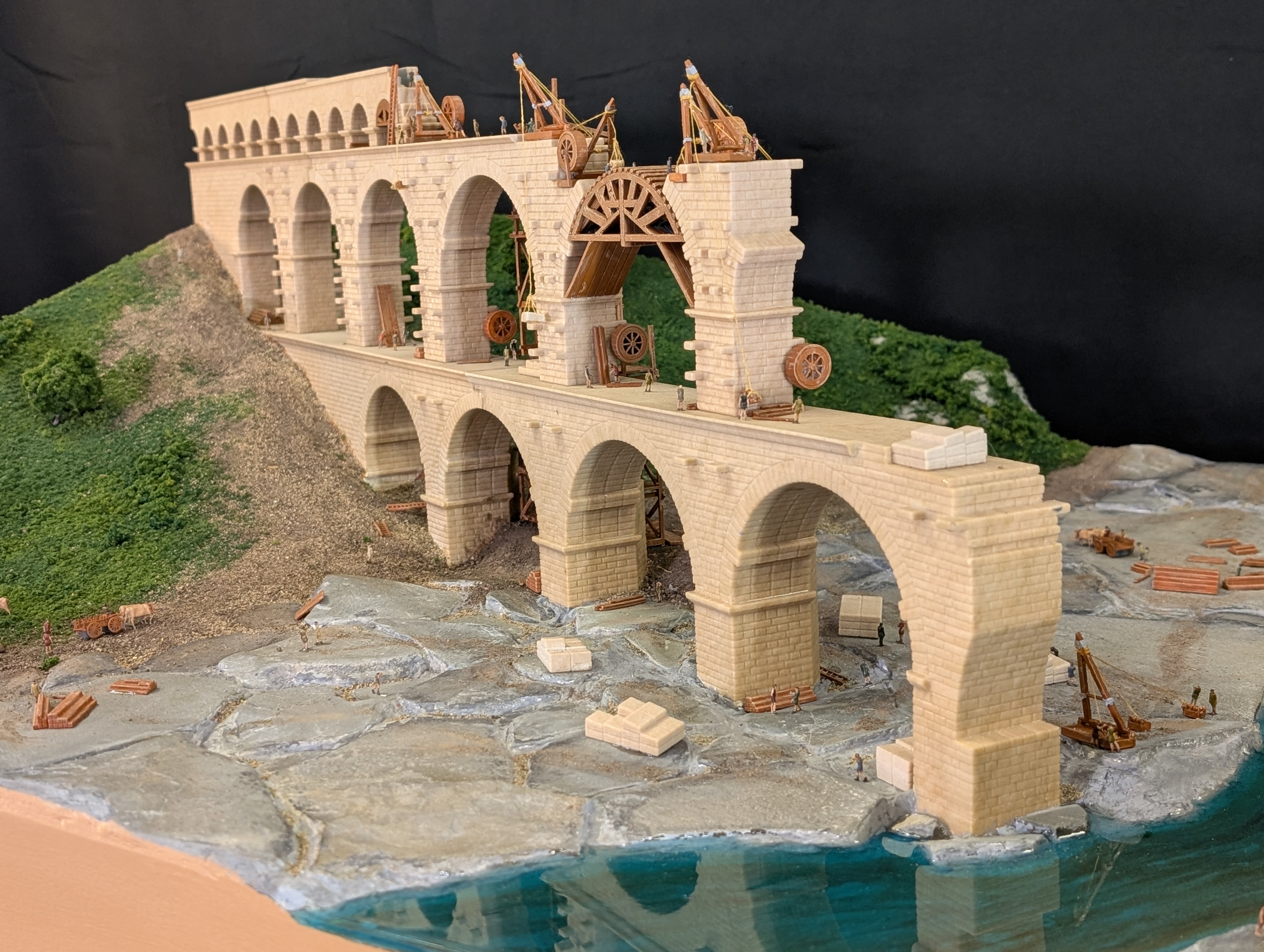 Scale model of a stone aqueduct bridge under construction with miniature workers and cranes, set beside a green hill and water.