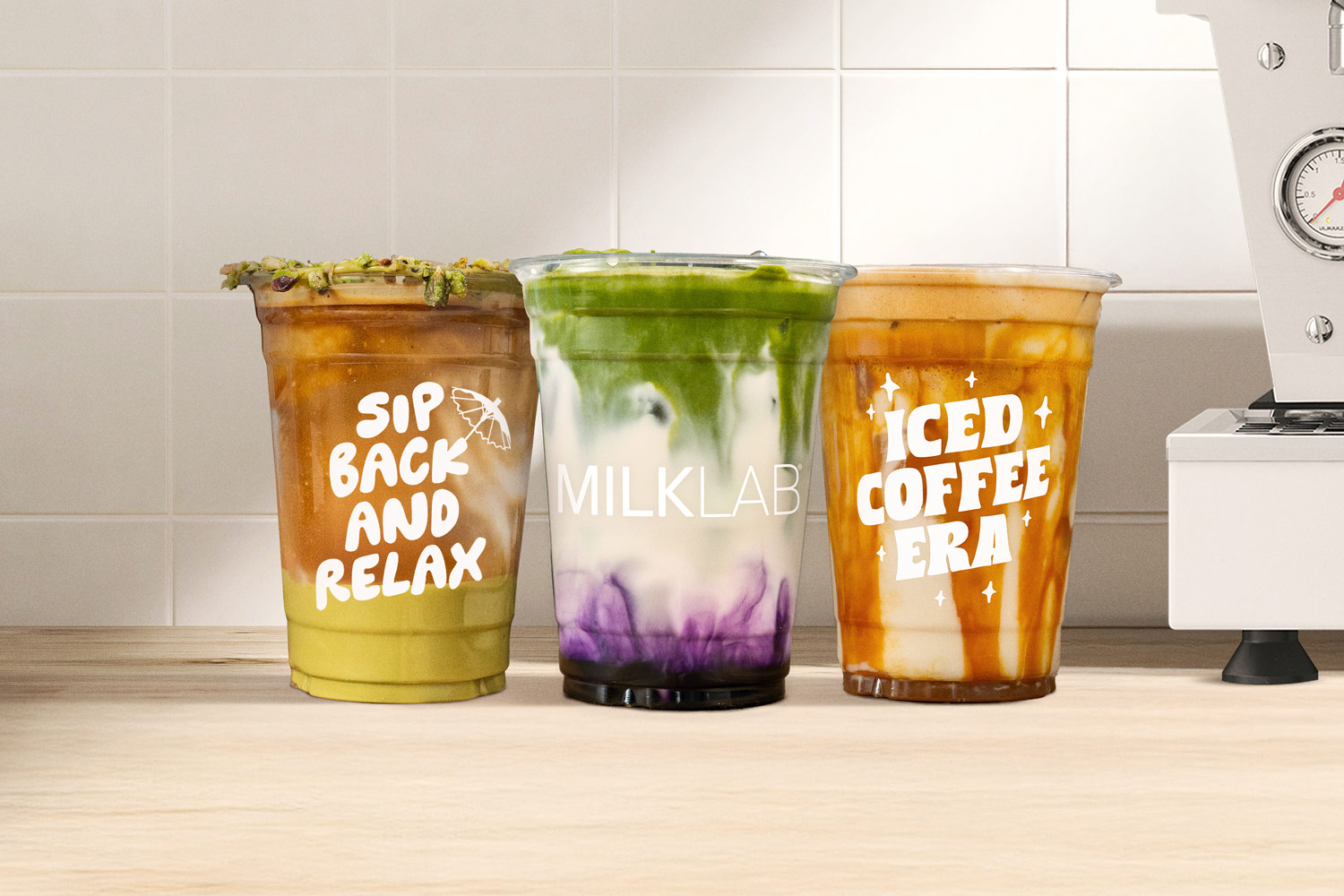Three colorful iced drinks on a wooden counter in clear plastic cups labeled 'Sip Back and Relax,' 'MILKLAB,' and 'Iced Coffee Era,' with a tiled wall and part of a coffee machine in the background.