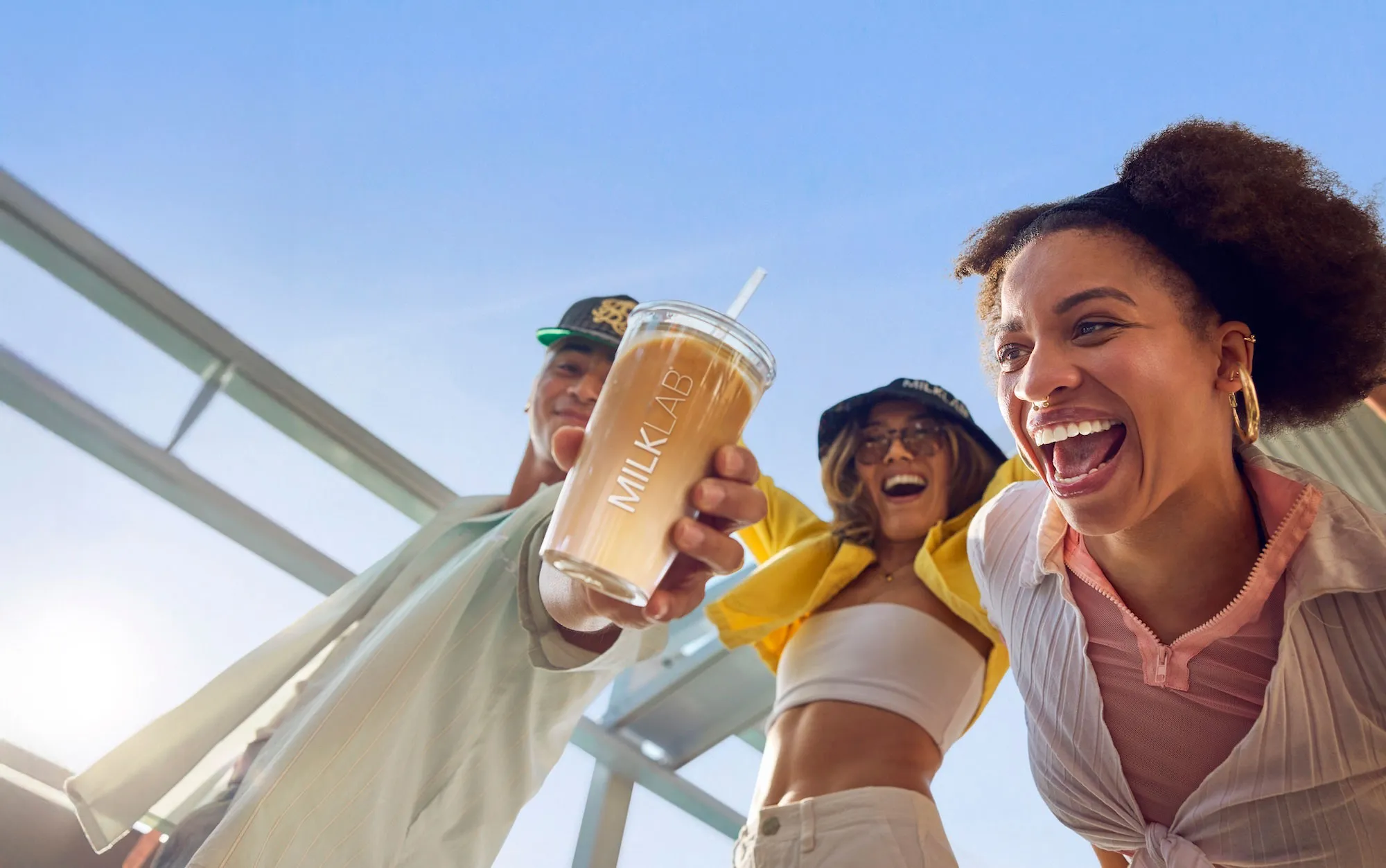 Three friends laughing outdoors as one holds a cup of iced coffee with the MilkLab logo against a clear blue sky.